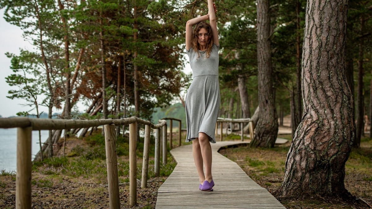 A slim dark-haired woman in a gray dress walking along a wooden boardwalk by the sea, surrounded by coastal forest.