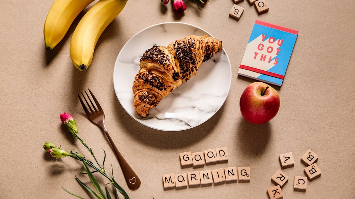 A croissant on a marble plate with a gold fork, apple, bananas, roses, You Got This notebook and Good Morning scrabble tiles