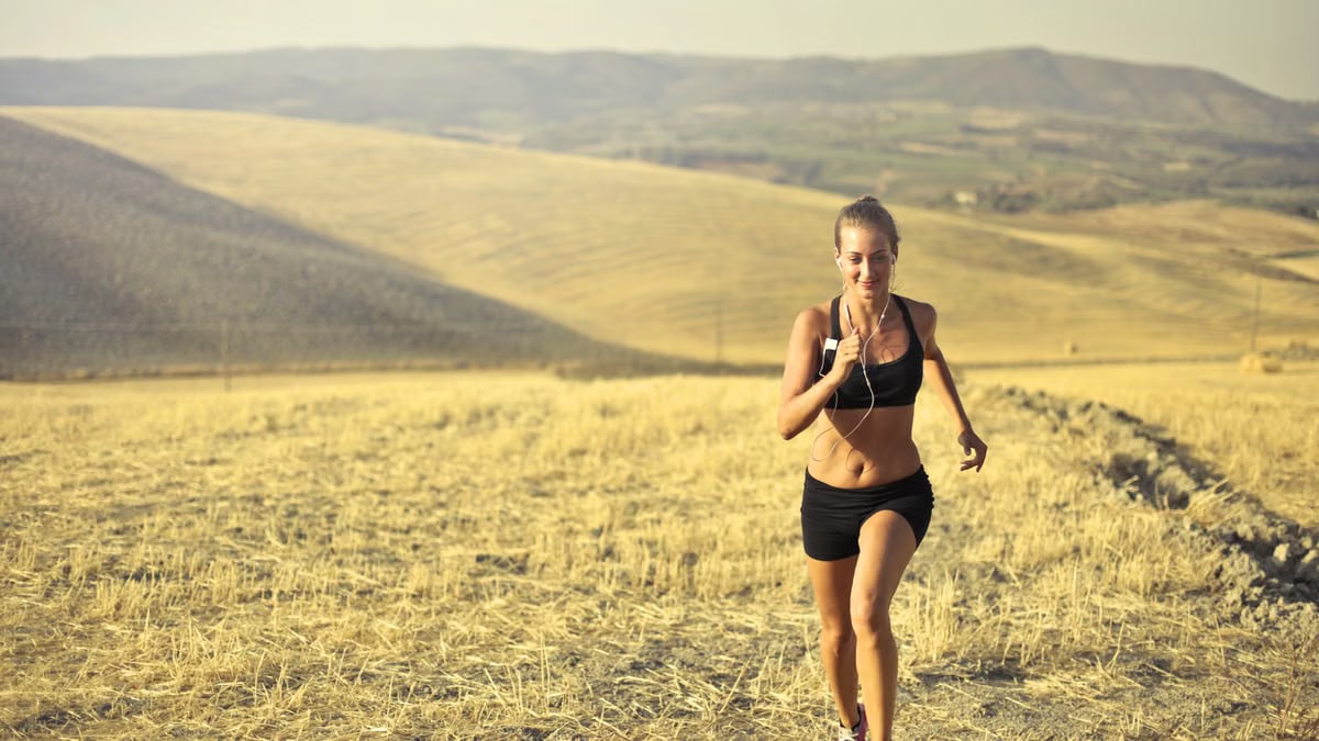 Powerful young female athlete in activewear running along hill on background of mountainous landscape and listening to music in earphones during cardio training
